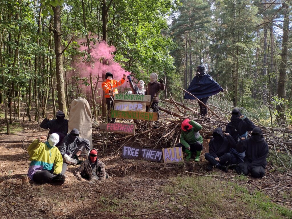 Zu sehen sind 13 Menschen, die vor bzw hinter Schilder stehen. Auf den Schilder steht ,Solidarität mit der unfreiwilligen Feuerwehr, free them all!'. Im Hintergrund brennt ein pinker Bengalo