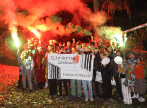 Menschen mit Transparent "Klimakrise löschen - Knäste abfackeln" vor rotem Rauch, teilweise in Halloween-Kostümen